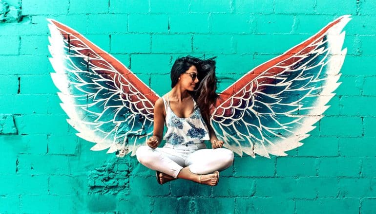 A young woman in white pants and a flower-covered top jumps in front of angel wings painted on a brick wall so that it looks like she's levitating while in a cross-legged sitting position