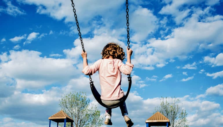 A young girl in pink swings at a park, with a cloudy blue sky in front of her and her back to the camera