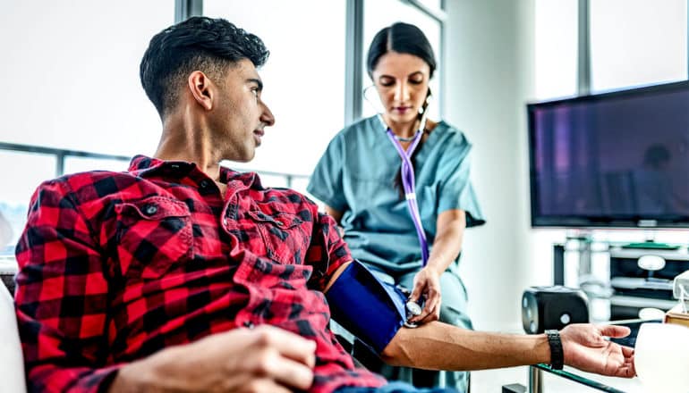 A man in a red and black checked shirt gets his blood pressure taken while seated at a doctor's office