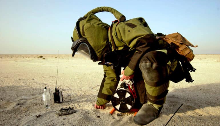 person in bomb suit and hood kneels in desert