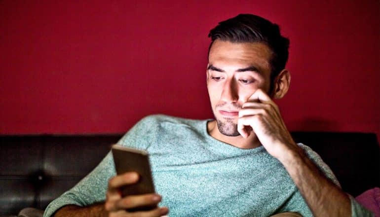 A man in a blue/green shirt watches a video on his phone while sitting on a leather couch with a red wall behind it