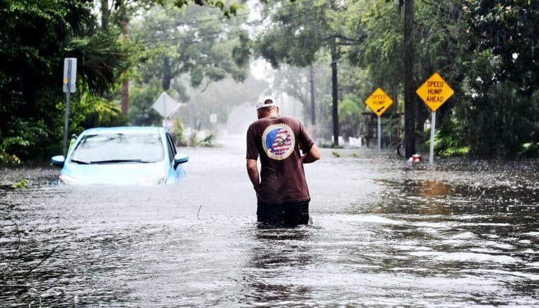 A man in dark pants, a brown t-shirt, and a white hat walks through flooded streets with water up to his waist. A blue, half-submerged car stands to his left