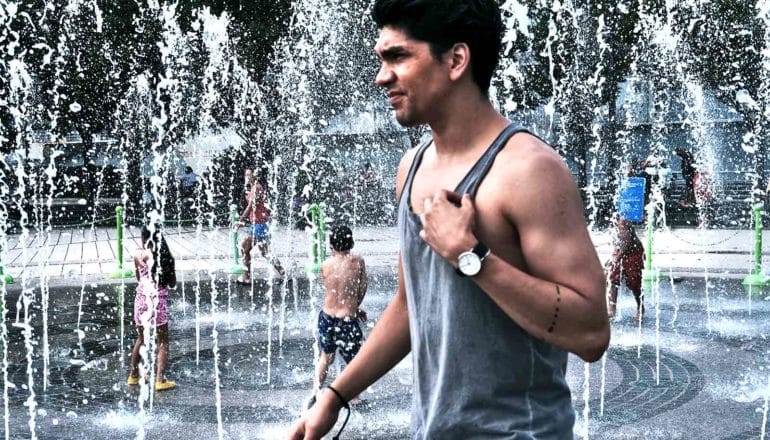 A man in a tank top walks past a city fountain where children are playing. He looks uncomfortable in the summer heat