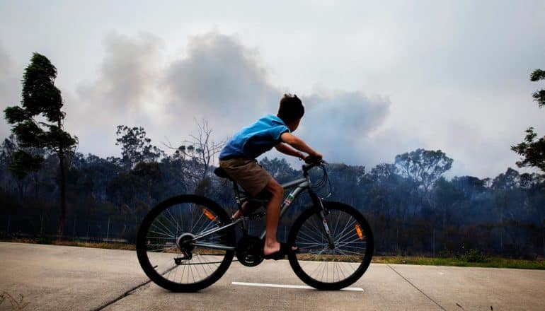 child rides bike with smoke from wildfire behind in the distance