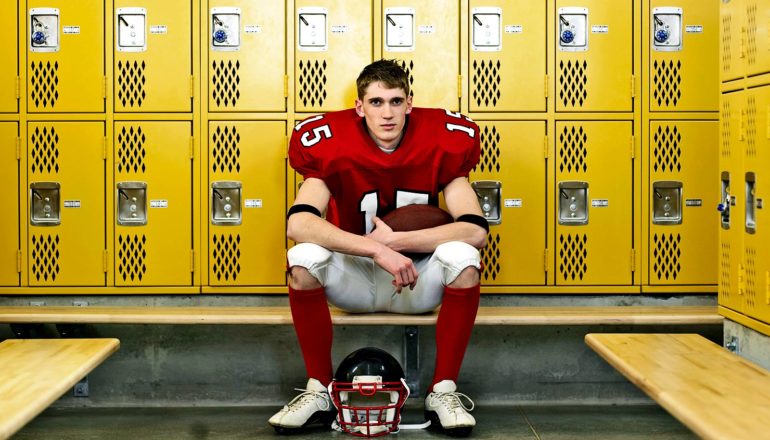 A young football player sits in a locker room in uniform with his helmet off