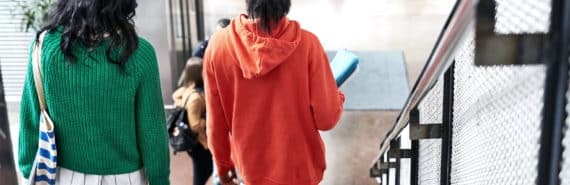 Two college students walk together down some stairs in a student union, one wearing a green sweater, the other wearing an orange hoodie