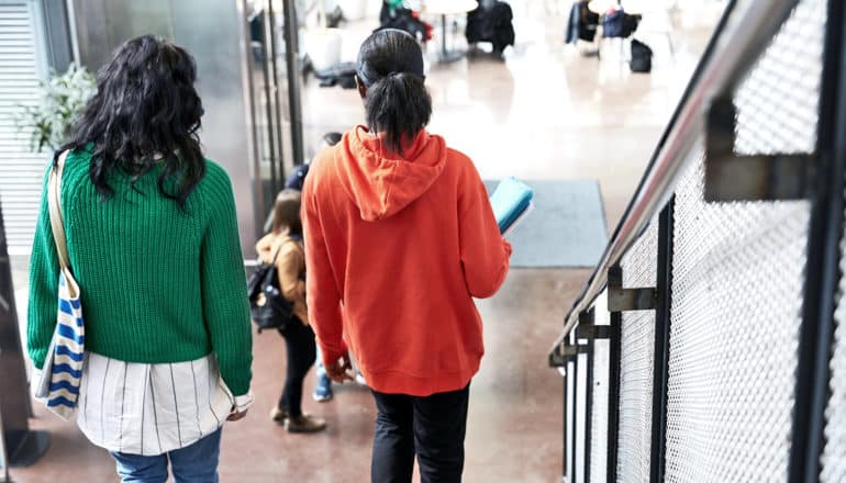 Two college students walk together down some stairs in a student union, one wearing a green sweater, the other wearing an orange hoodie