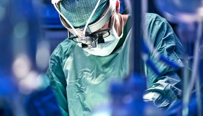 A surgeon in green scrubs and a white mask looks down while performing surgery