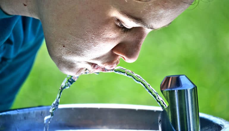 A young woman leans over a water fountain to take a drink as the light reflecting off the fountain's metal illuminates her face, with a deep green background