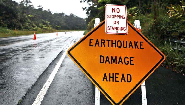 An orange sign on a road surrounded by trees reads "Earthquake Damage Ahead"