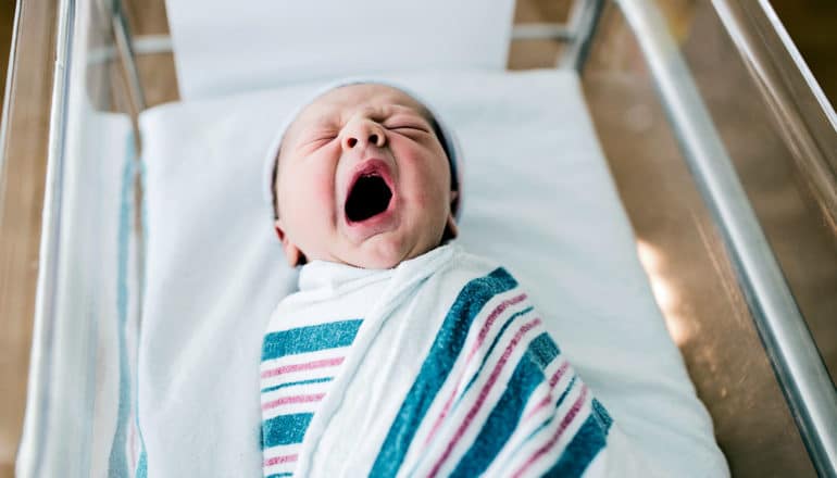 A newborn baby is yawning while wrapped in a white blanket with colored stripes in a hospital bassinet