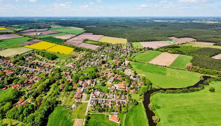 An aerial shot shows farm fields, a small town, and forests near a river, with the sky in the far background