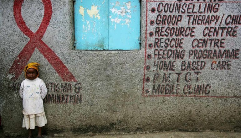 child stands against wall with sign that lists "counselling, group therapy, resource centre, feeding programme, home based care, pmtct, mobile clinic"