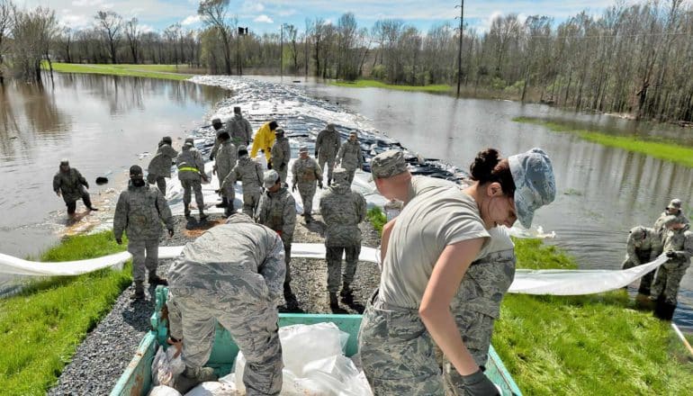 people in camo outfits move sandbags on levee between floodwaters