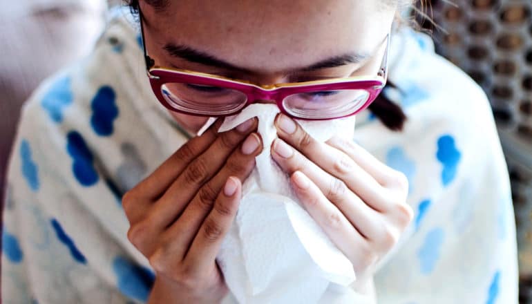 A young woman blows her nose with a tissue as she sits wrapped in a blanket in a chair