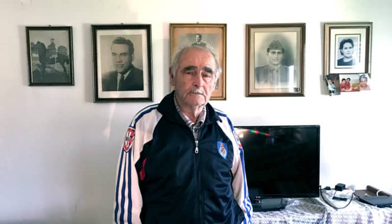 older man stands in front of framed photos (horse, young man in uniorm)