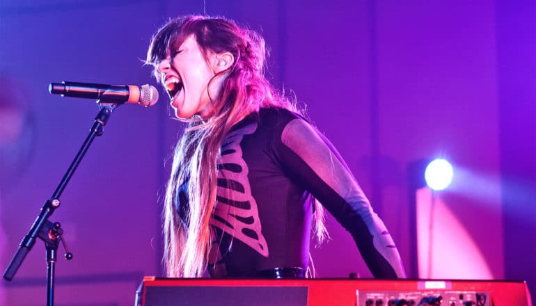 A singer in a skeleton shirt shouts into a microphone while on stage under purple lights