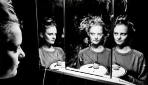 A young woman sits in front of a three-mirror vanity, her reflection appearing in each mirror at a different angle, with incense burning on the small table in front of her