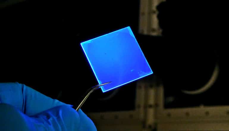 A researcher wearing a blue glove holds a pair of tweezers that are holding a pane of glass that's emitting intense blue light