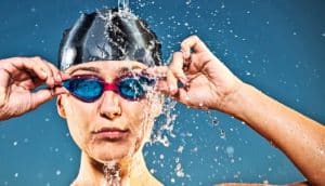 Water splashes down on the head of a swimmer wearing pink goggles with blue lenses and a black cap while she reaches up to adjust her goggles