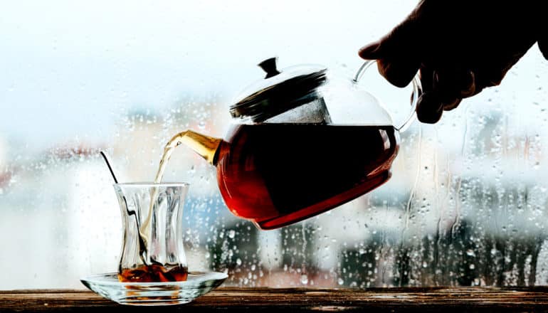 A hand holds a tea pot and pours it into a glass cup with a spoon in it against a rain-covered window in the background