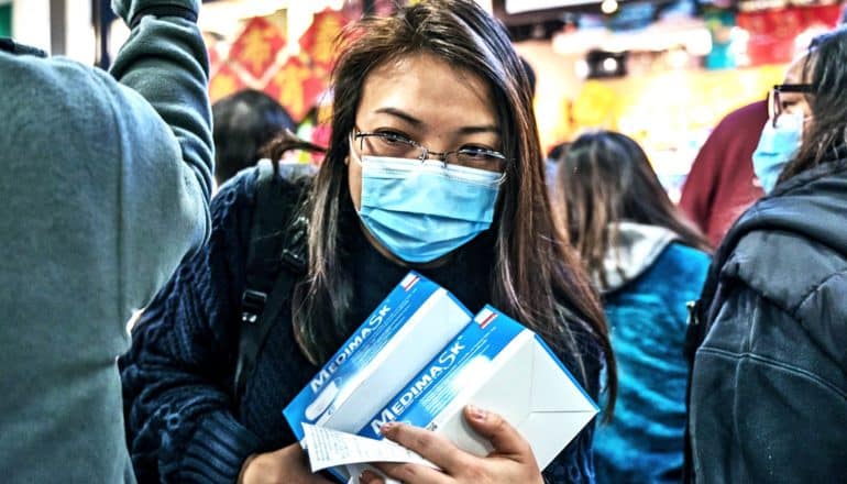 A woman wearing a blue medical masks carries two boxes of more medical masks as she moves through a crowd