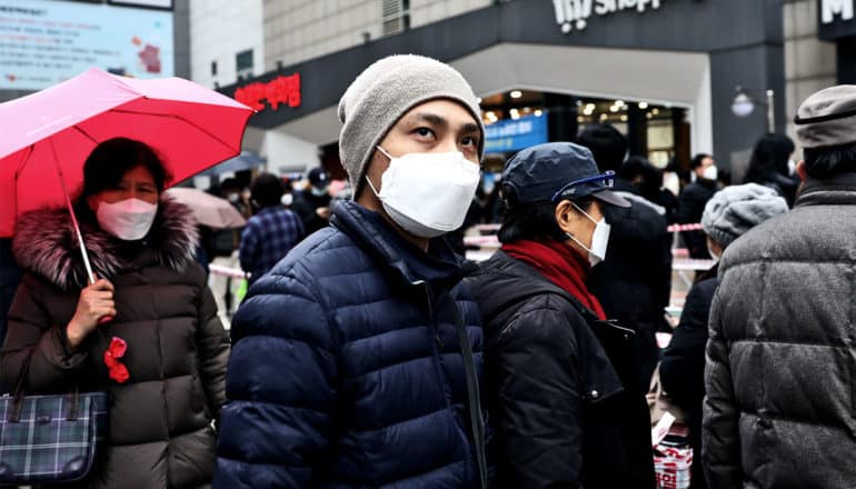 A man in a medical mask, parka, and beanie looks up while walking on a busy street, where other people are also wearing medical masks