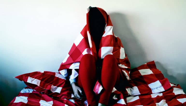 A woman has wrapped herself in a red and white checked comforter while sitting on their bed, so that her face is obscured