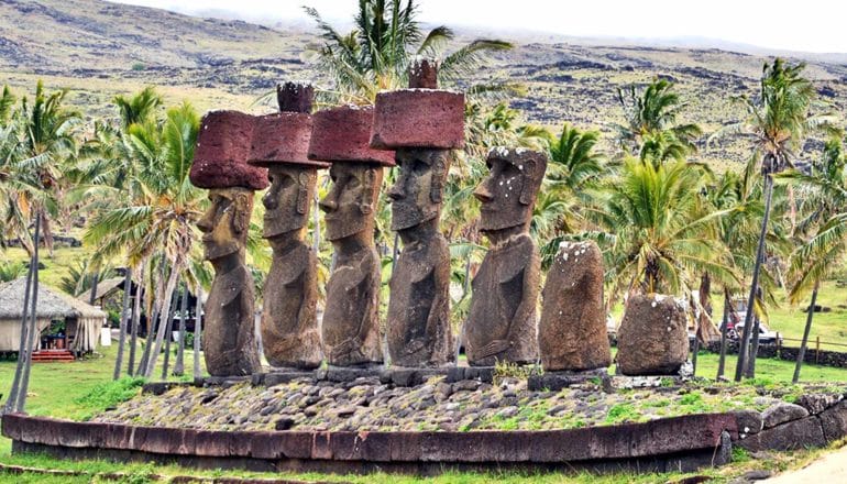 Statues on Easter Island stand in a row, some decayed and fallen apart, with palm trees and hills in the background