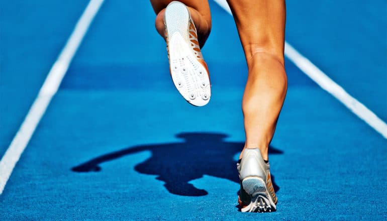 A runner has one foot on the blue track and one in the air, with her shadow visible against the track