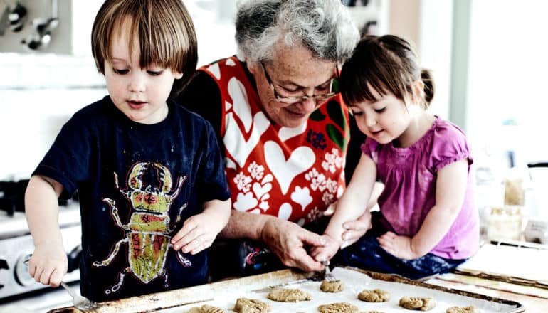 A grandmother makes cookies with her two grandchildren in a mostly-white kitchen