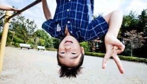 A young boy shows the peace sign with his right hand as he's hanging upside down from monkey bars at a park