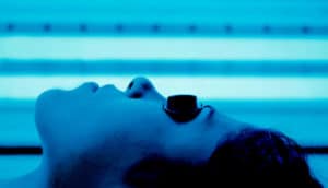 A woman lies in an indoor tanning bed, with her bathed in blue light, while wearing black goggles over her eyes