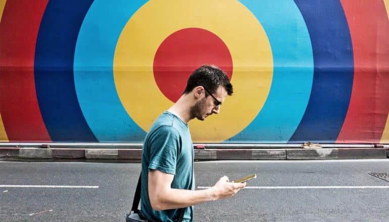 A man walks down the street looking down at his phone. The wall behind him is a multi-colored target design that's centered behind his head