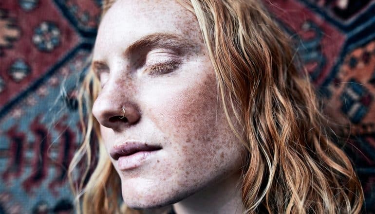 A woman with a nose ring closes her eyes with a tarp or wall rug in the background