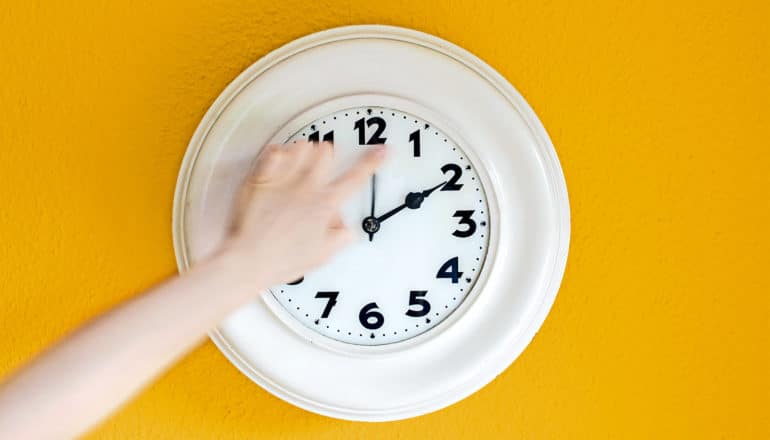 A hand reaches out from off-camera to turn back a white clock on a yellow wall