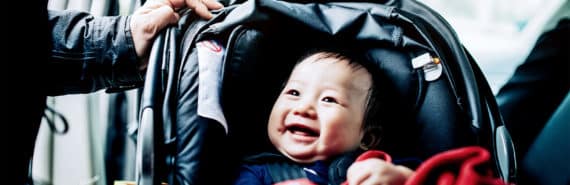 A mother talks to her baby, who's sitting in a car seat looking up at her and smiling, covered with a red blanket