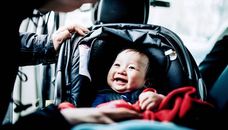 A mother talks to her baby, who's sitting in a car seat looking up at her and smiling, covered with a red blanket