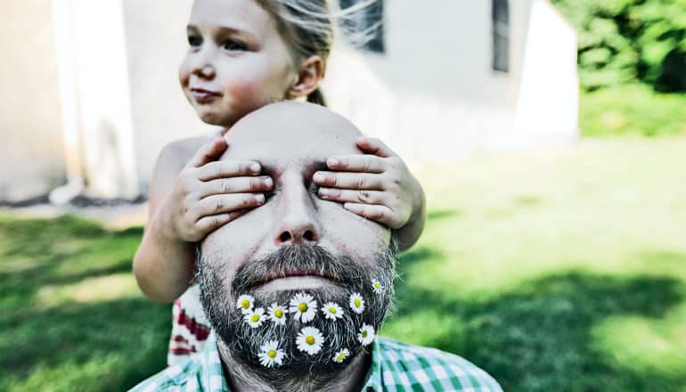 A bearded man has his eyes covered by a little girl behind him, with his beard full of flowers and a grassy lawn and white house behind them.