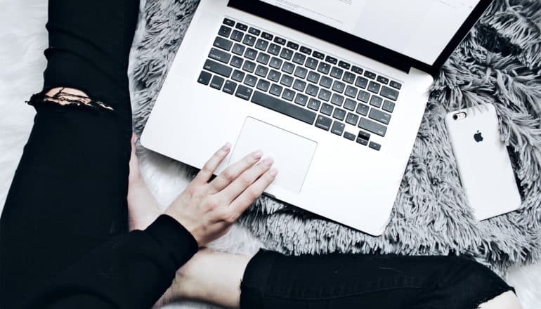 A woman works on her laptop with her white phone next to her on a gray pillow, with her torn black jeans in the frame