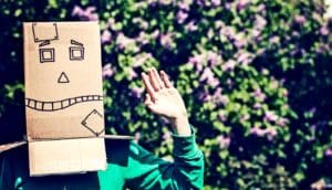 A person wearing a robot mask made out of a cardboard box on their head waves to the camera, with leaves and flowers in the background