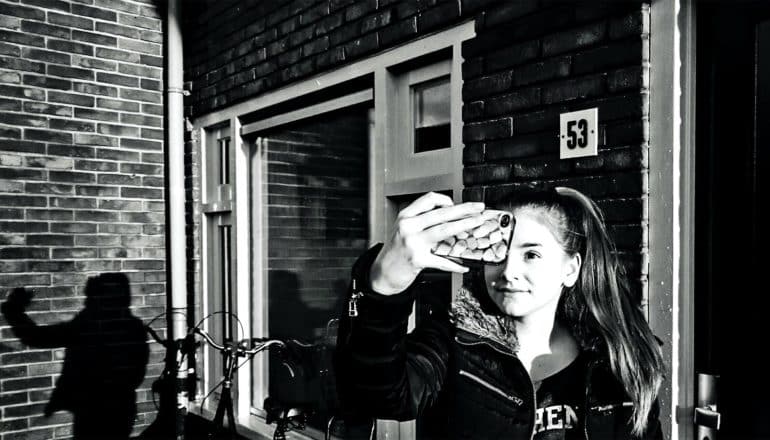 A teen girl takes a selfie in front of a brick building, her shadow appearing on the wall to her right