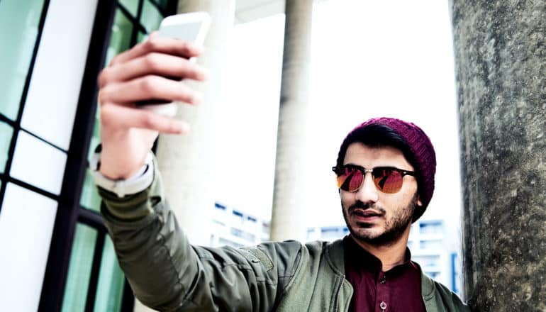 A young man in a green jacket, dark red polo shirt, sunglasses, and a beanie takes a selfie