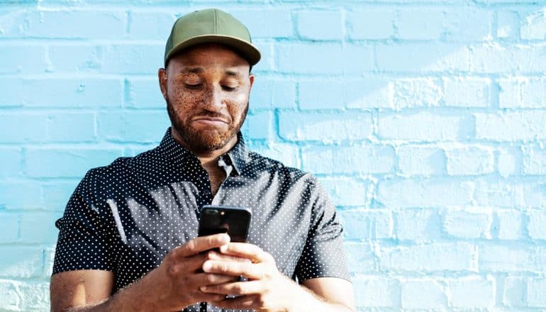 A man in a black and white polka-dot shirt and green baseball cap looks down at his phone while making a skeptical face, while standing against a brick wall painted blue