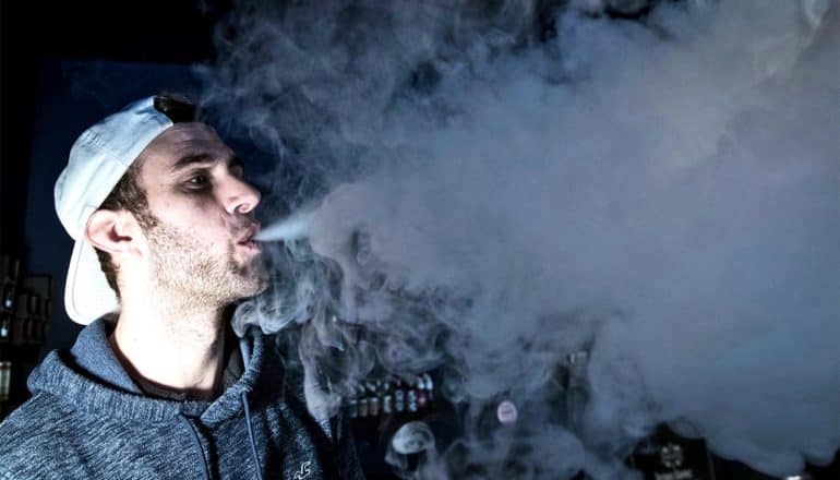 A man in a white cap and gray hoodie exhales vapor from an e-cigarette