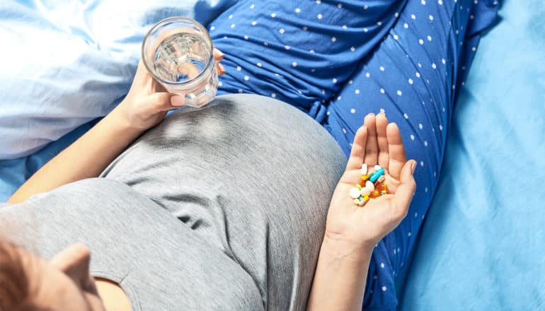 A pregnant woman in blue and gray pajamas sits on her bed holding a glass of water and a handful of vitamins.