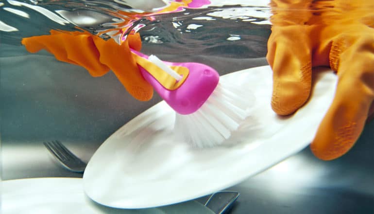 A person wearing orange gloves scrubs a white plate with a pink brush in a sink full of water