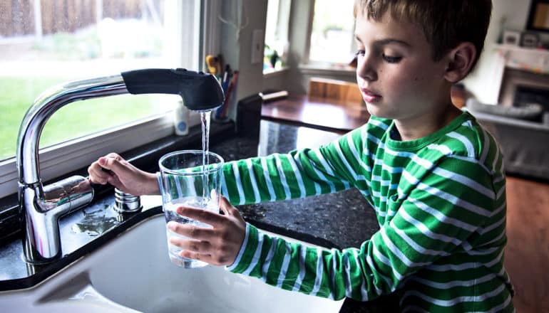 A young boy in a green long-sleeved t-shirt pours a glass of water from the kitchen faucet