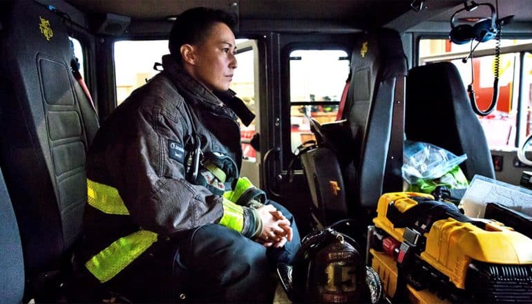 A woman in firefighting gear sits in a fire truck looking out the windshield with her hands folded