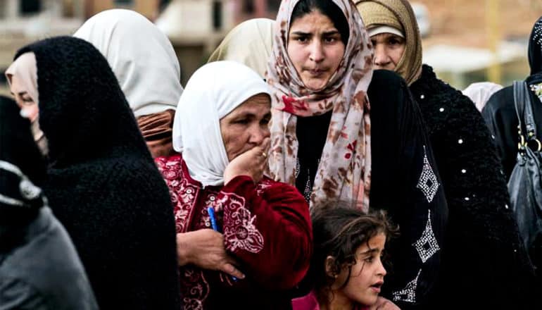 A line of refugee women look deeply concerned. A young girl stands with them, looking off to her left.